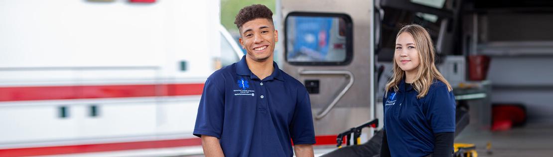 两个EMT学生在一辆救护车后面微笑着摆姿势